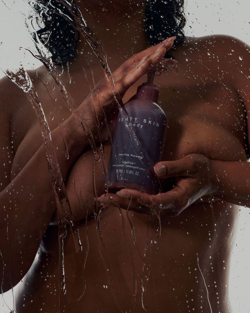 An adult woman holds a bottle of Fenty Skin body product in a beauty product shot with water droplets on the lens.