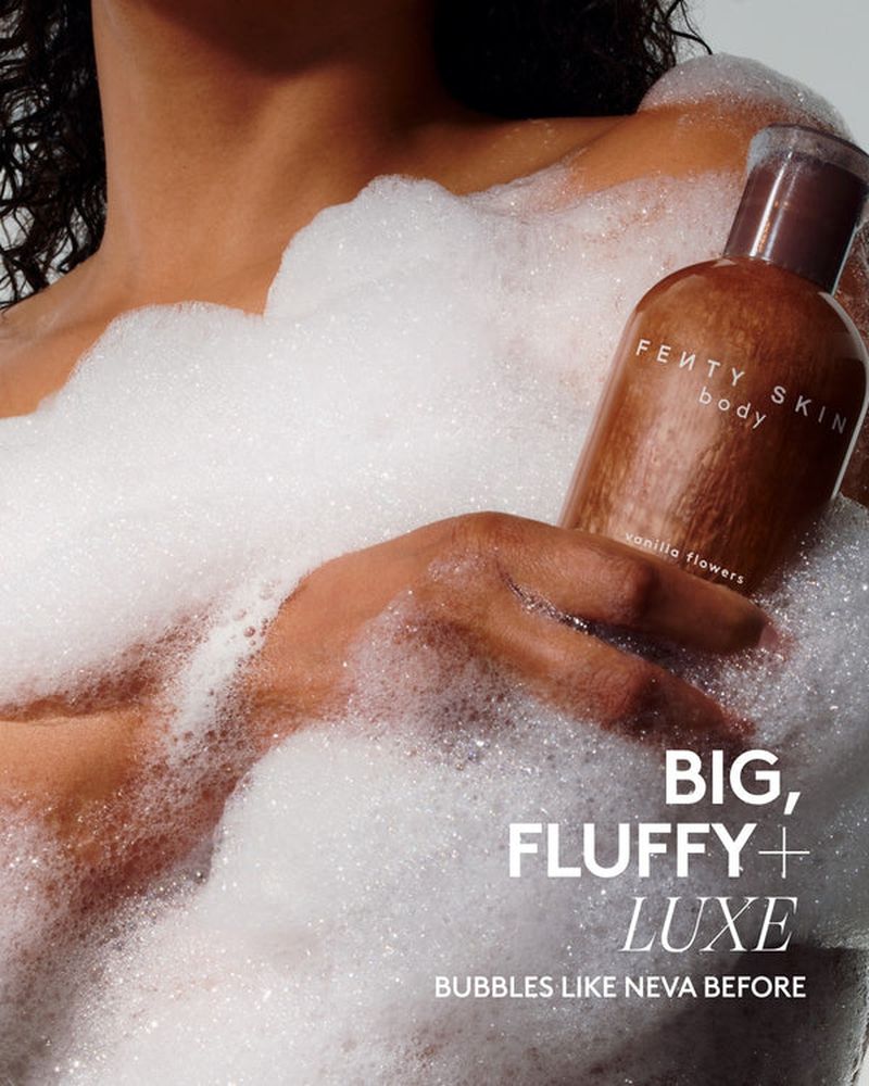Tan-skinned model holds a Fenty Skin body wash bottle surrounded by fluffy white bath bubbles, studio lighting, tan, brown, and white.