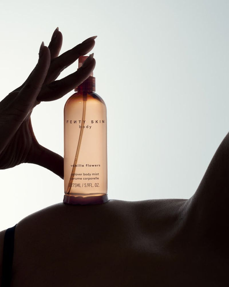 Fenty Skin body mist spray bottle held by a manicured hand, resting on a woman's shoulder in a beauty and skincare product shot.