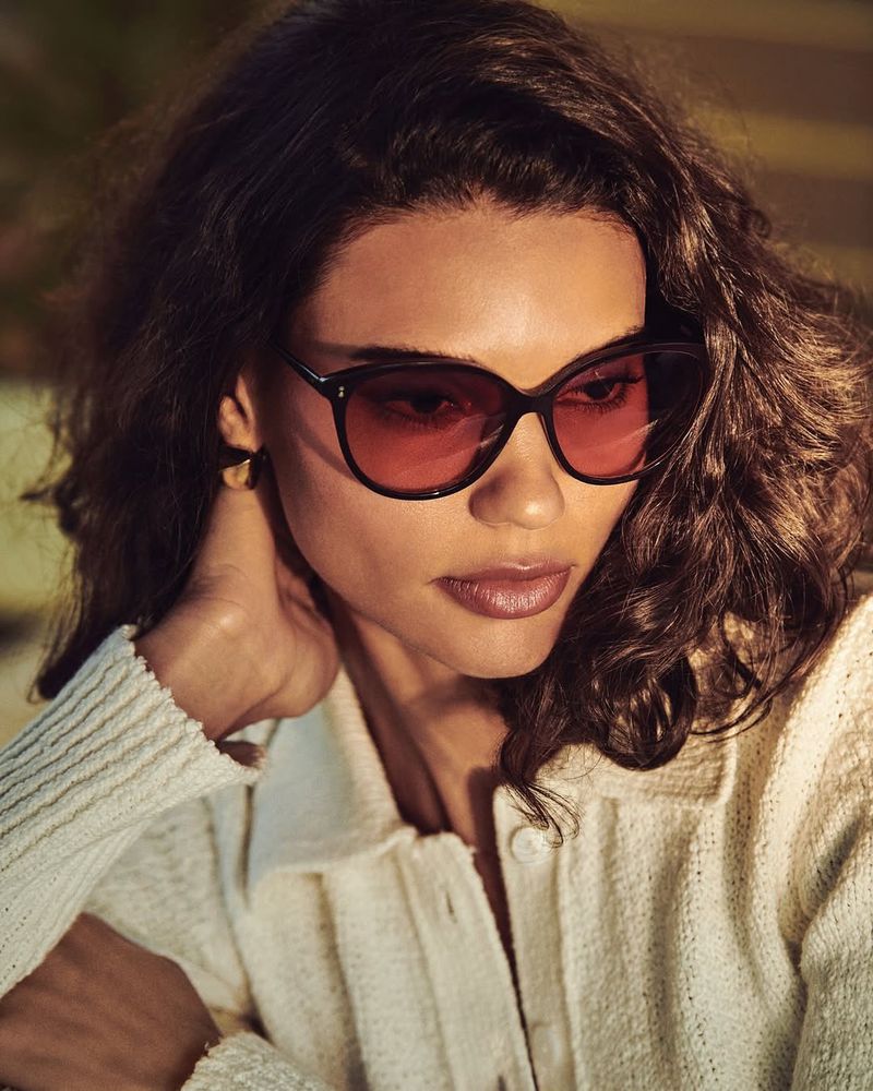 Woman with dark curly hair wearing sunglasses and a white button-up shirt in a close-up indoor portrait.