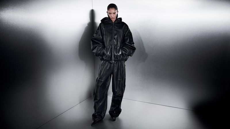 A person in a hooded leather jacket and matching pants stands in front of a gray corner wall, bathed in studio lighting for a fashion shoot.