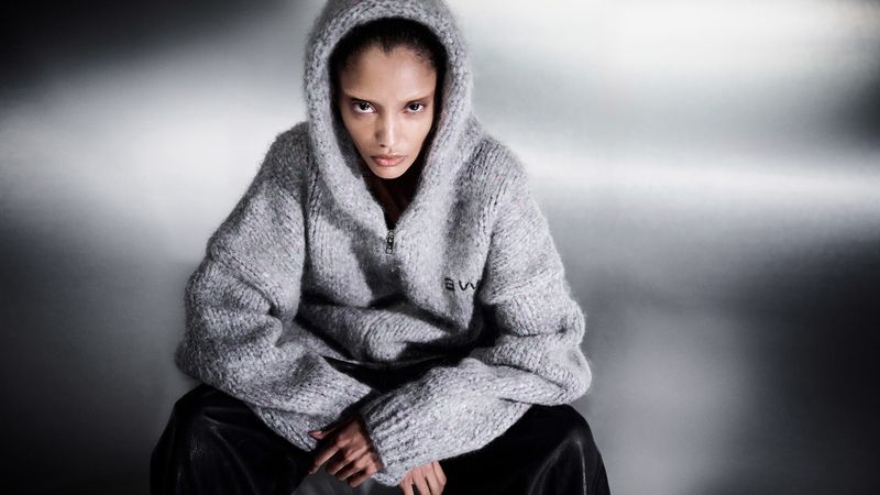 Adult female model in a hooded grey knit sweater and black pants sitting against a textured grey background in a medium shot, fashion.