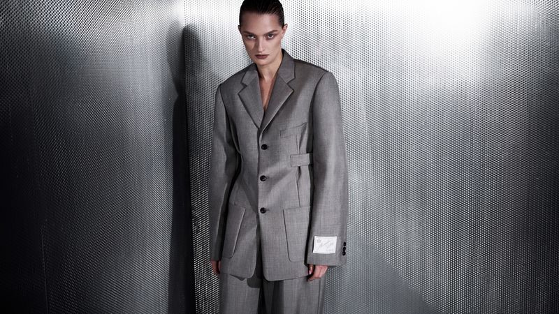 Fashion shot of a model in a gray oversized suit, looking at the camera, against a textured metal wall in a photo studio.