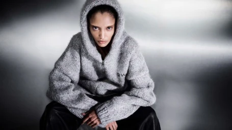 Model wearing an oversized grey knit hooded sweater and black leather pants against a reflective metallic backdrop.