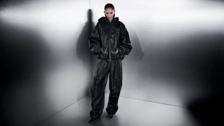 Model wearing an oversized black leather hooded jacket and matching wide-leg leather trousers against a brushed metal corner backdrop.