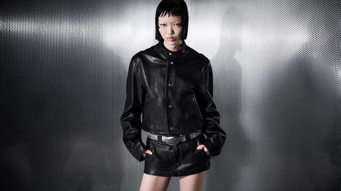Model Weiyi Fang wearing a black leather jacket and skirt with a metallic belt against a perforated metal backdrop.