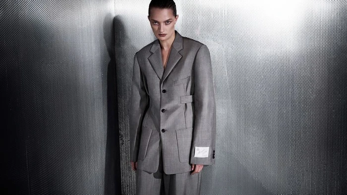 Model in a grey tailored suit jacket posing against a perforated metal wall background