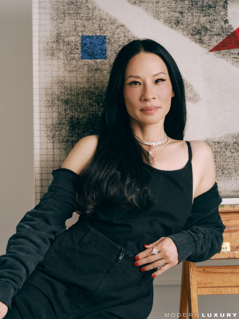 Portrait of Lucy Liu sitting with a black outfit and elegant jewelry in front of an abstract patterned wall. Bright, neutral lighting.