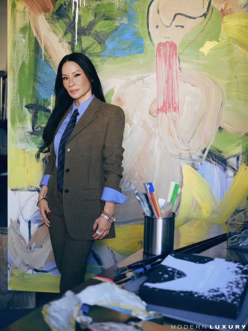 Lucy Liu posing in a brown suit jacket with an abstract painting, in front of a desk, with visible art supplies in the setting.