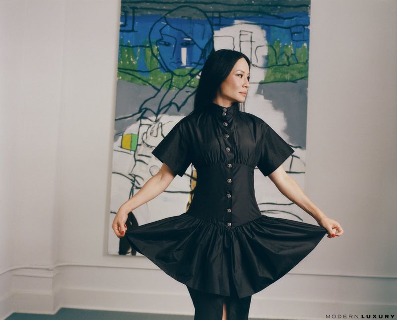 Lucy Liu poses in a black dress against an abstract painting in a studio. The image showcases high fashion editorial style.