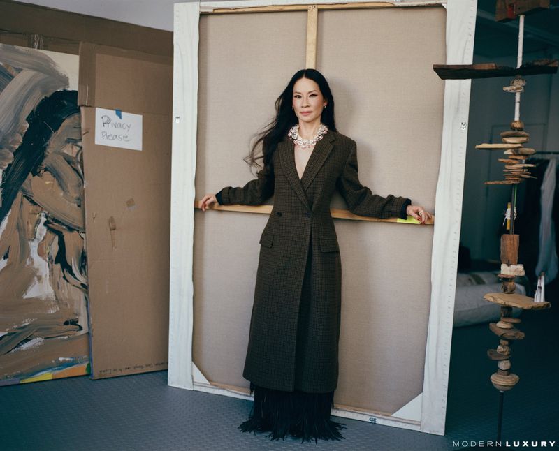 Lucy Liu leans against a canvas in a photo studio wearing a long brown coat, fringed skirt, and pearl jewelry, with art and studio props.