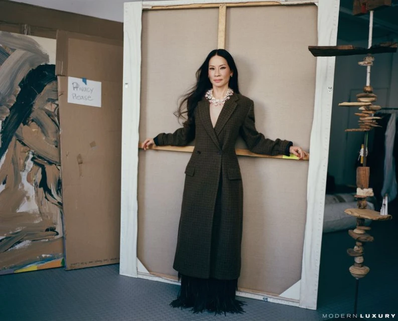 Lucy Liu leans against a canvas in a photo studio wearing a long brown coat, fringed skirt, and pearl jewelry, with art and studio props.