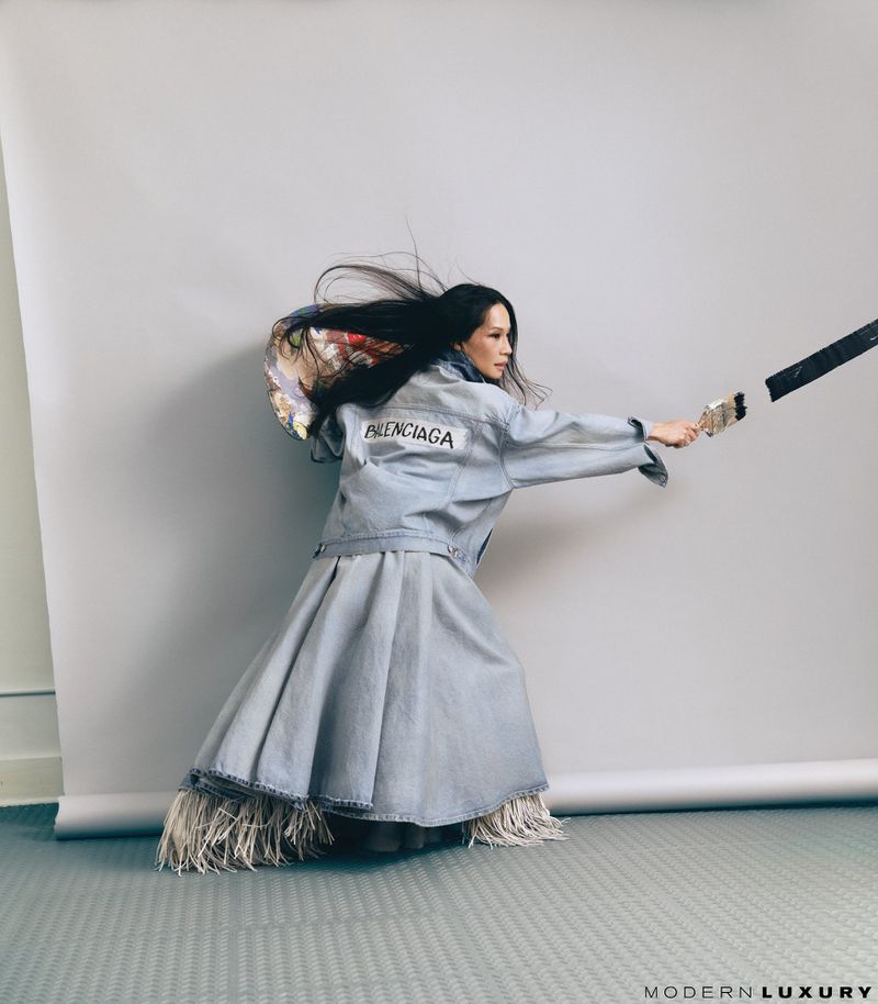 Woman in denim jacket and skirt holds paint brush, posing in front of grey background in a fashion photography studio setting.