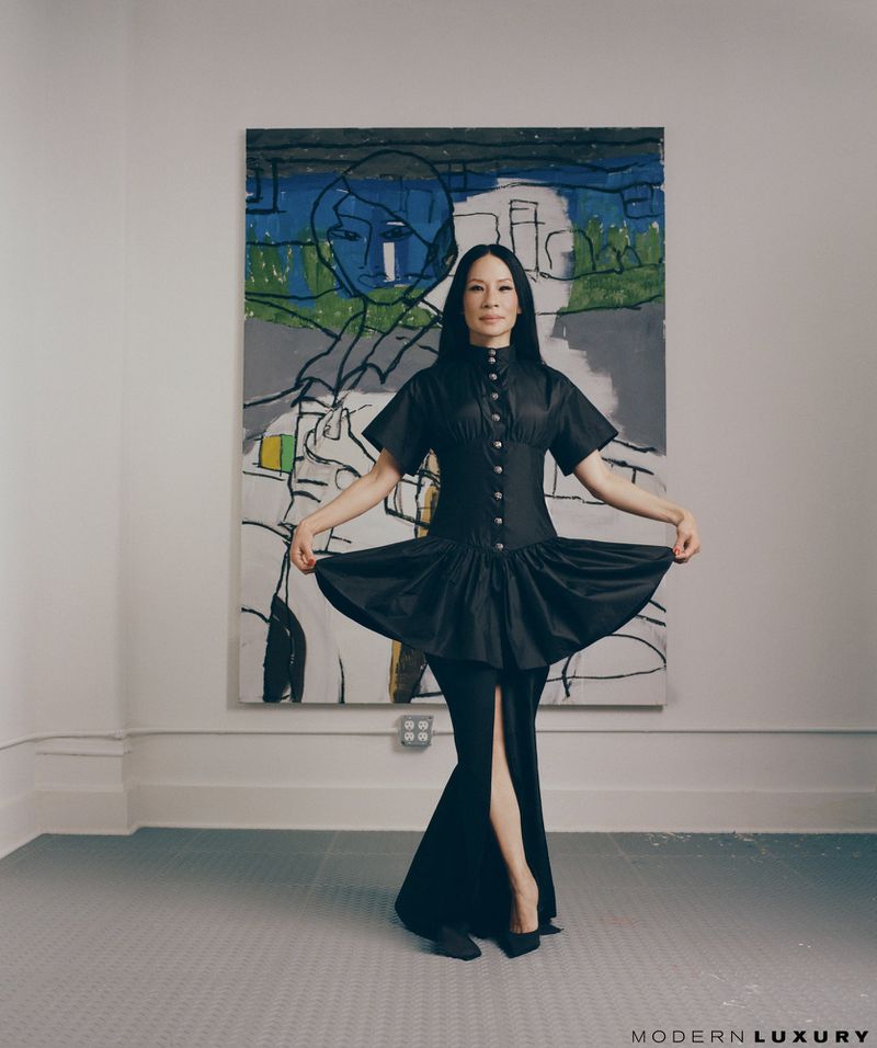 Portrait of an adult woman wearing a black dress posing in front of a colourful painting in a studio with a grey tile floor.