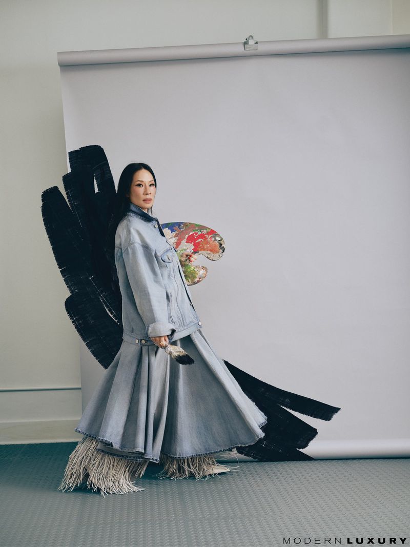 Stylish woman in denim jacket and layered skirt posing with art palette and brush in a studio setting with black brushstroke backdrop.