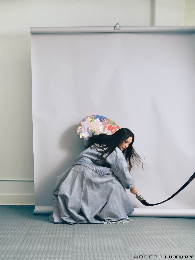 A woman in a denim outfit sits near a white backdrop, appearing to paint with a large brush in an indoor photo studio setting.