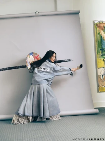 Woman in denim fashion painting with a brush against a gray backdrop, wearing a Balenciaga jacket and a skirt with fringe.