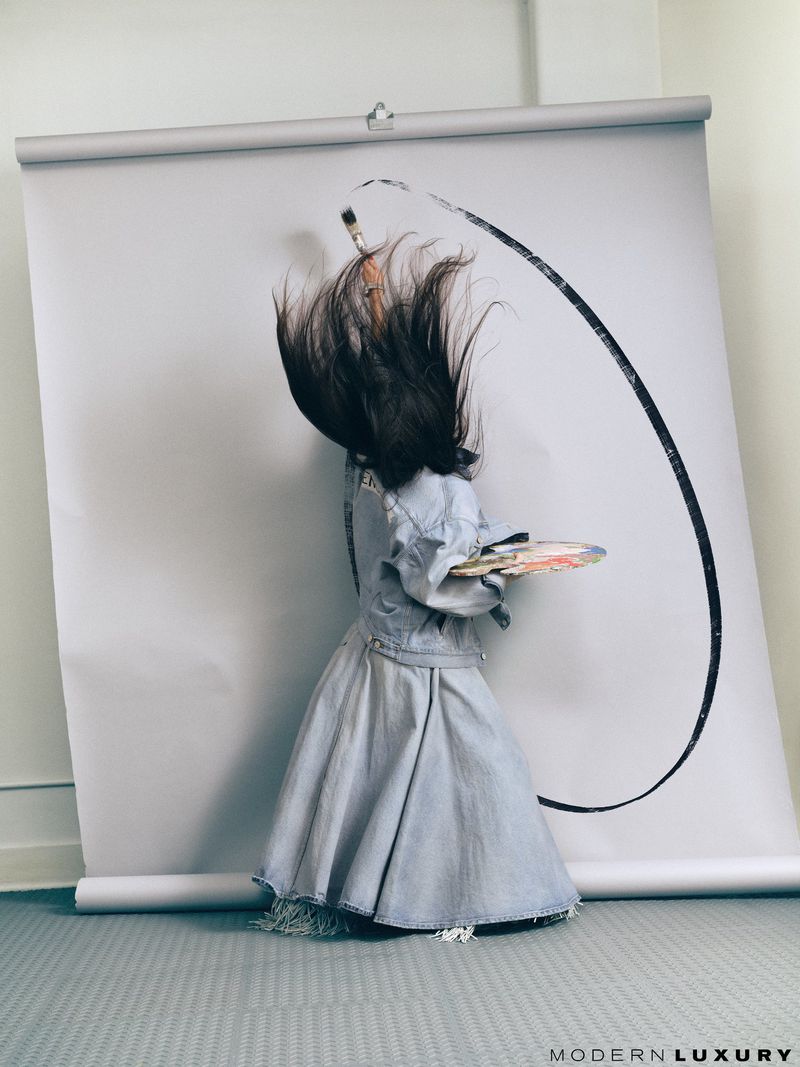 Person in denim dress holds paint palette standing in front of a white backdrop with a paintbrush and black paint stroke. 