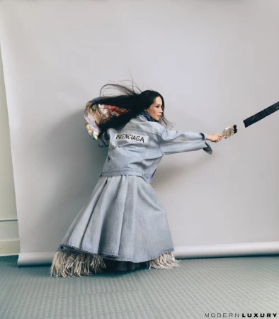 Lucy Liu in a Balenciaga denim jacket and matching flared skirt holding a paintbrush in a studio setting.