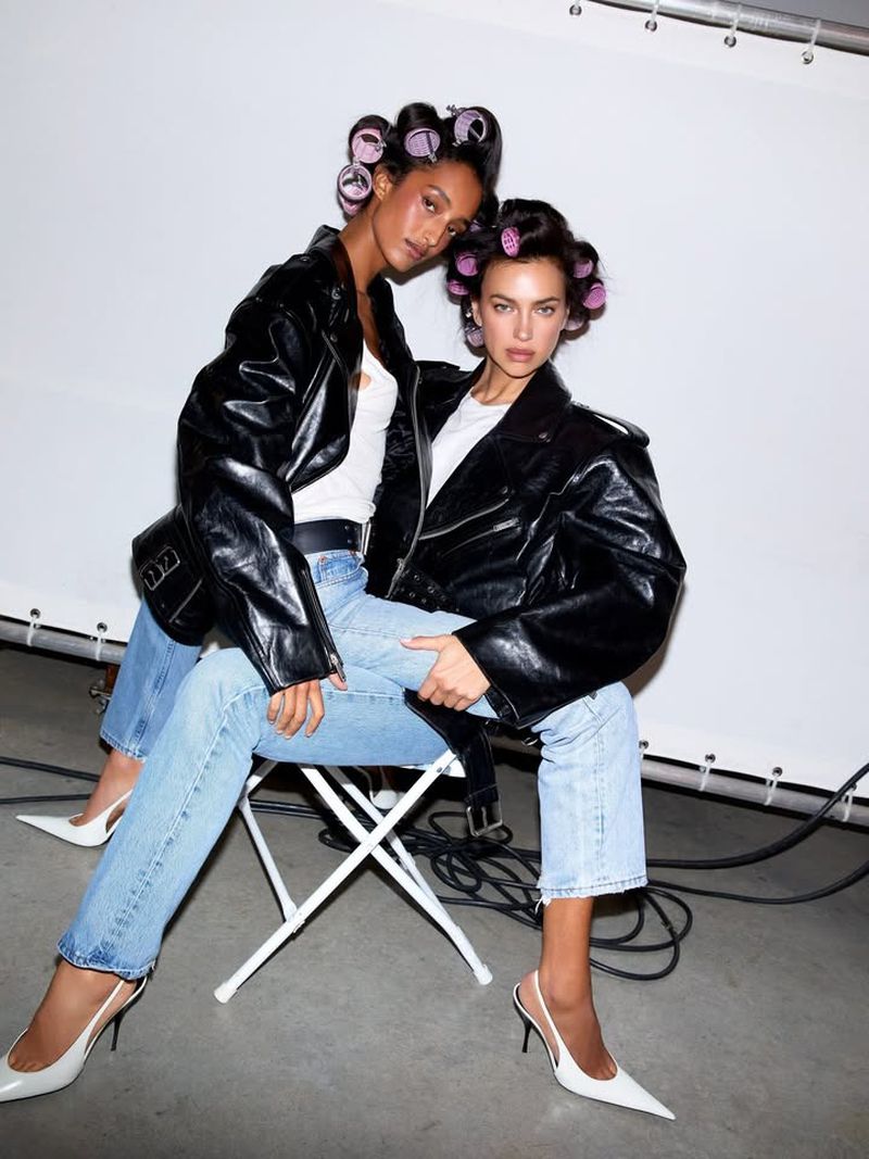 Two women pose in a photo studio wearing black leather jackets, light-wash jeans, white heels, and hair rollers against a white backdrop.