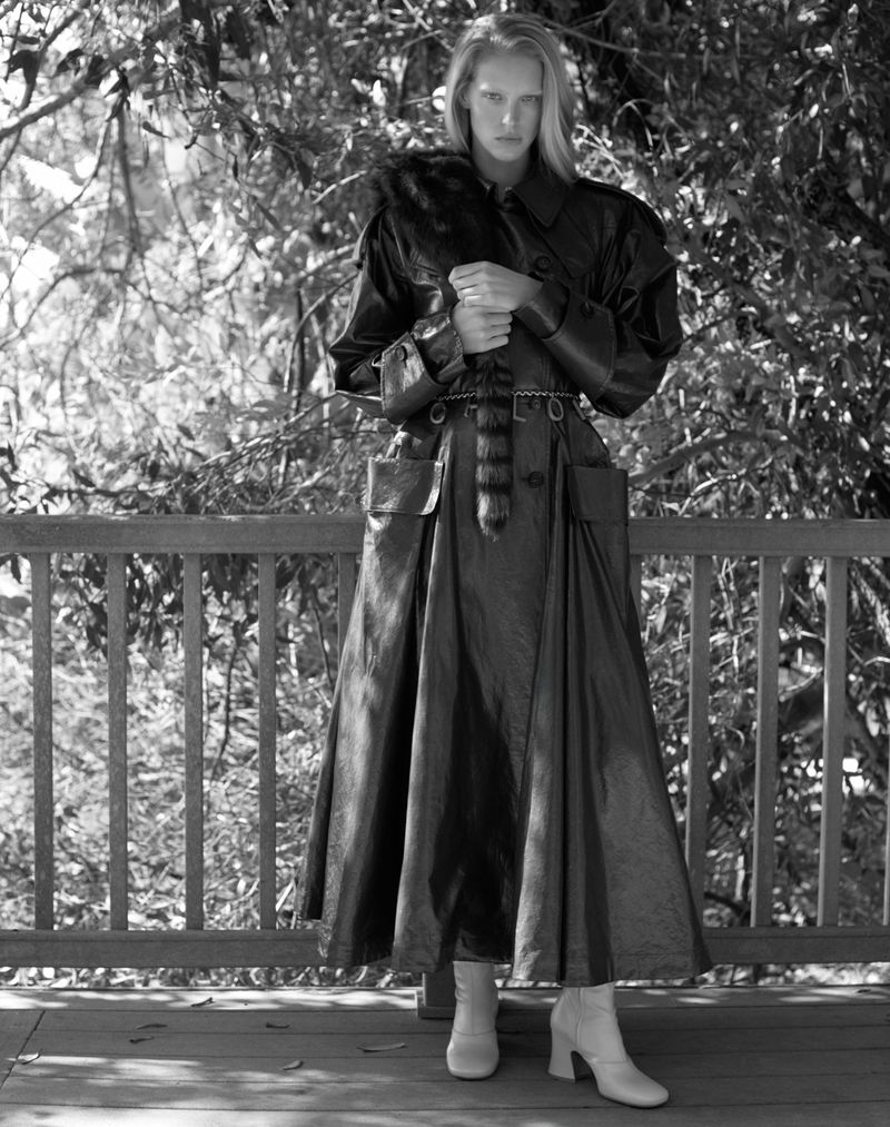 Black and white fashion shot of a tall, slender woman in a dark, shiny trench coat, outdoors on a wooden deck with a forest background.