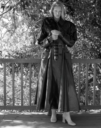 Black and white fashion shot of a tall, slender woman in a dark, shiny trench coat, outdoors on a wooden deck with a forest background.