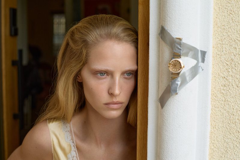 Close-up of a blonde woman near a door frame with a gold watch taped to the wall; neutral expression, indoor setting, natural light.