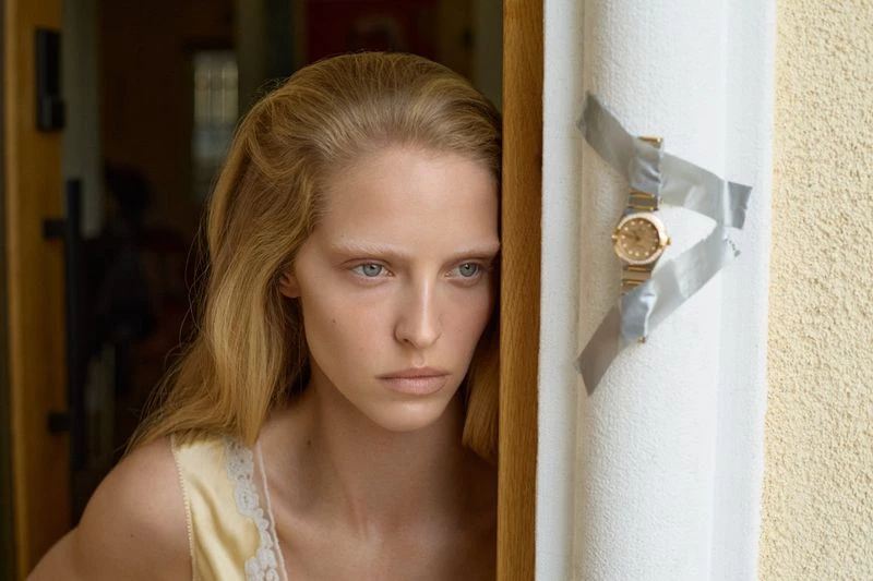 Close-up of a blonde woman near a door frame with a gold watch taped to the wall; neutral expression, indoor setting, natural light.