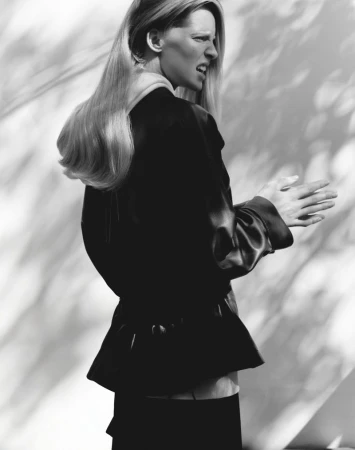 Monochrome studio portrait of a blonde adult female, wearing a dark jacket, with dappled lighting effects creating shadows on the background.