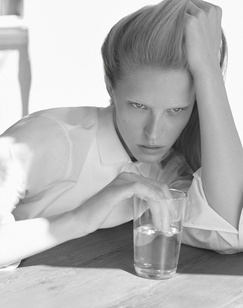 Black and white portrait of a fair-skinned adult woman with her hand on her head sitting at a table with a glass of water.