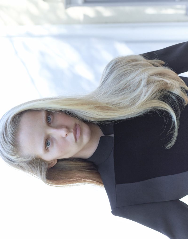 Model Abby Champion with long blonde hair wearing a high-neck black jacket against a bright diffused background.
