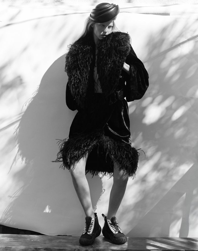 Monochrome fashion shot: woman in dark feather-trimmed coat, turban, and platform sneakers poses against a sunlit wall with leaf shadows.