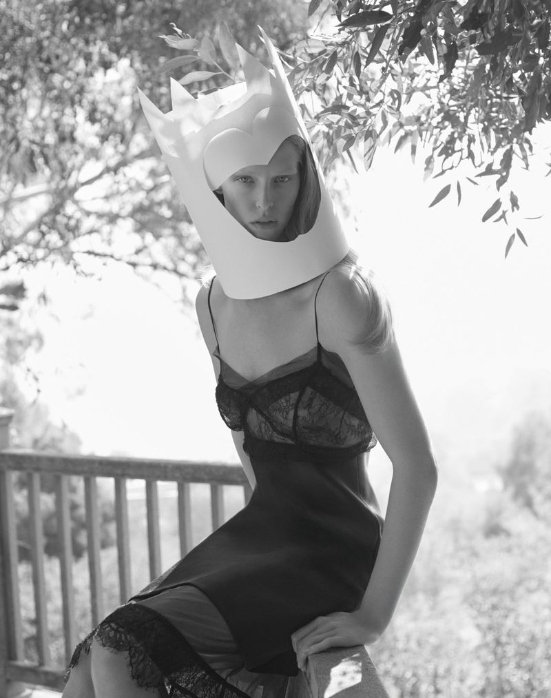 Black and white portrait of a woman in a black dress sitting outdoors with a white crown-like paper sculpture on her head.