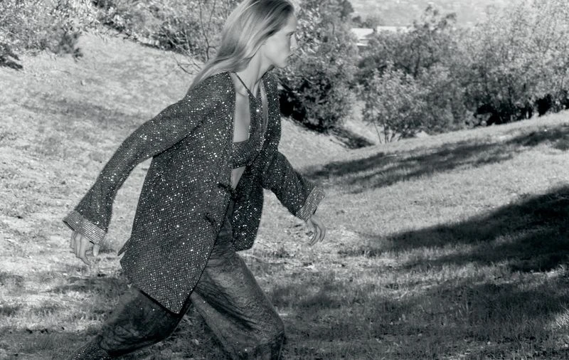 Black and white fashion shot of a blonde woman in a sparkling jacket and pants walking on a grassy hillside with trees in the background.