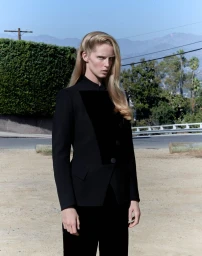 Model Abby Champion wearing a structured black tailored blazer outdoors against a roadside and hillside backdrop.