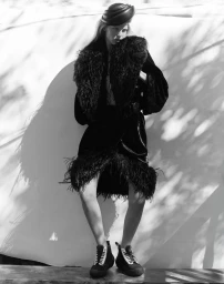 Model Abby Champion wearing a black feather-trimmed coat, sculpted hat, and high-top sneakers against a sun-dappled backdrop.