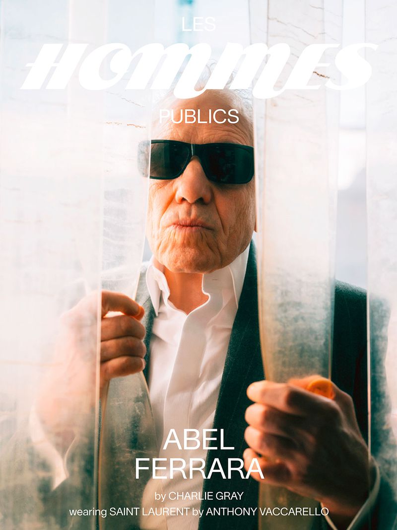 Abel Ferrara, photographed through a textured glass panel, wears sunglasses, a white collared shirt, and blazer in this celebrity portrait.