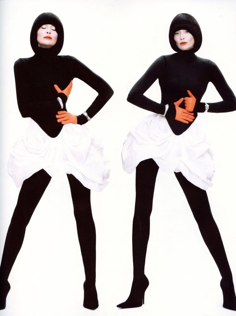 Two fashion models pose in black turtlenecks, white skirts, black tights, and heels against a stark white backdrop in a studio shot.