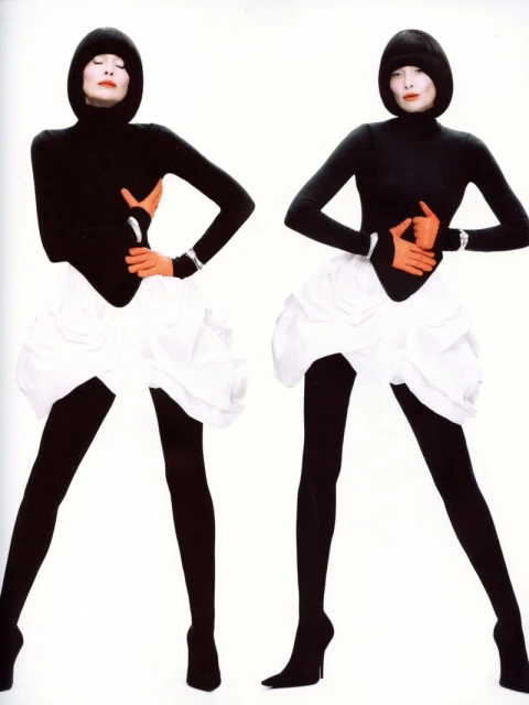 Two fashion models pose in black turtlenecks, white skirts, black tights, and heels against a stark white backdrop in a studio shot.