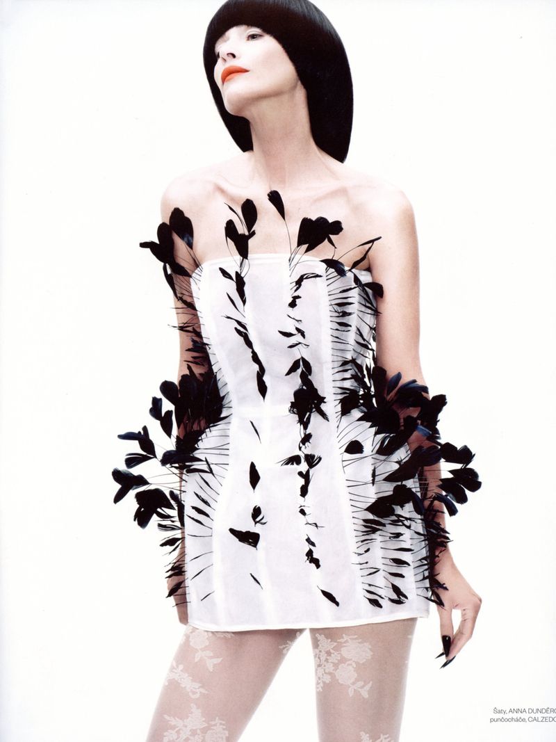 A slender woman with black bob haircut wears a white corset-style mini dress with black feather accents against a white background.