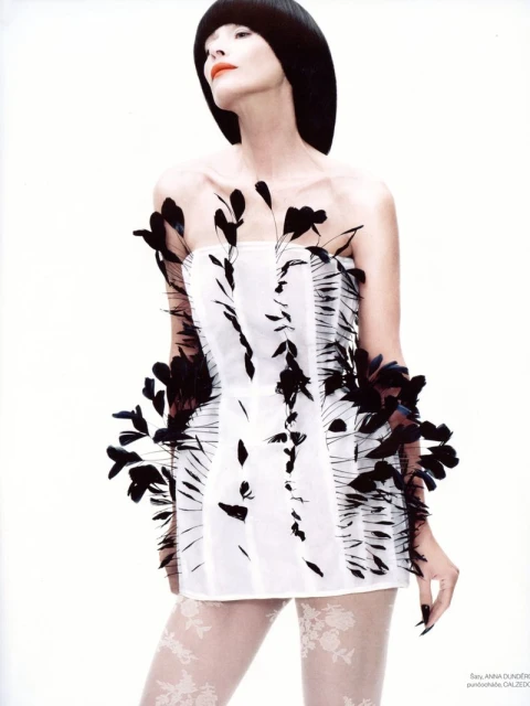 A slender woman with black bob haircut wears a white corset-style mini dress with black feather accents against a white background.