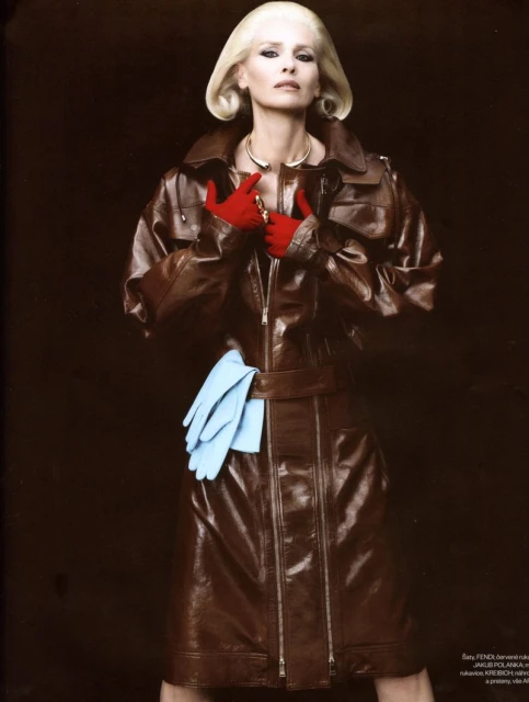 Fashion editorial of a blonde woman in a brown leather coat, with red gloves, accessorized with a gold necklace against a dark background.