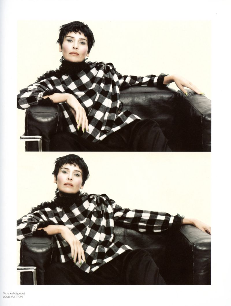 A woman in a black and white plaid shirt sits in a black leather chair against a plain backdrop in a fashion photography setting.