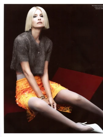 Blonde woman wearing a grey knit top and orange lace skirt seated on a red surface against a dark background for Vogue Czechoslovakia.