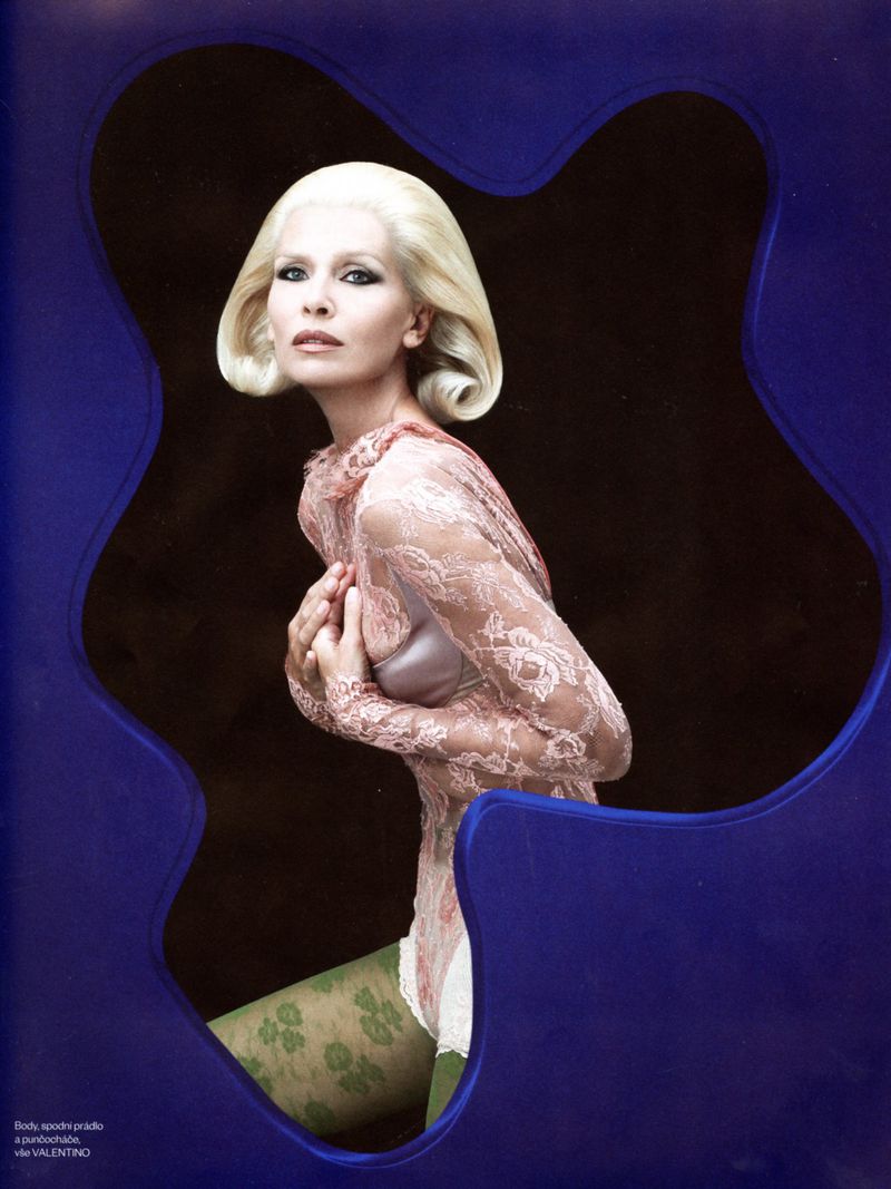 Elegant woman with platinum blonde wavy hair in a sheer lace top and patterned tights framed by a curved blue chair back.