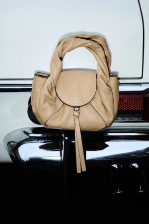 Elegant beige leather handbag with twisted top handle and front flap detail, resting on the chrome bumper of a white vintage car.