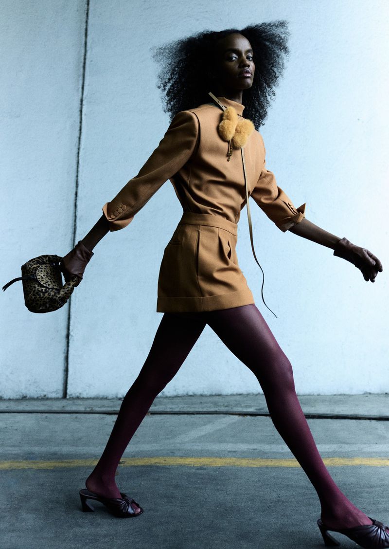 A slim, dark-skinned model in a tan suit and burgundy tights strides confidently with a patterned purse against a light gray backdrop.