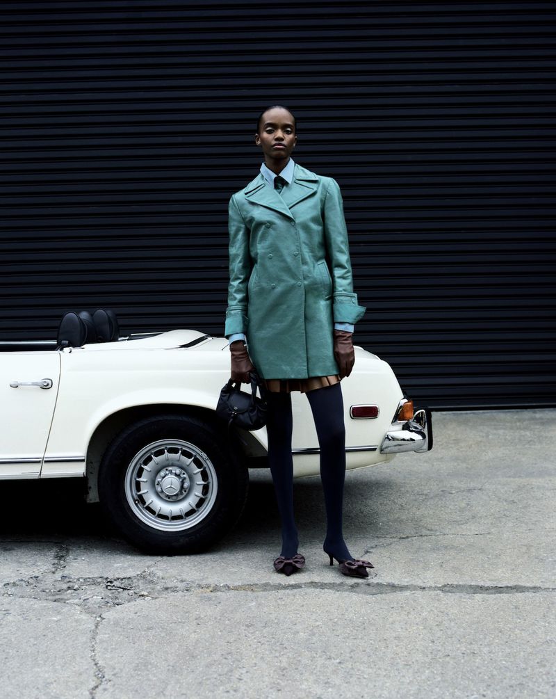 Model standing next to a vintage white convertible car in front of a dark corrugated wall for L'Officiel USA