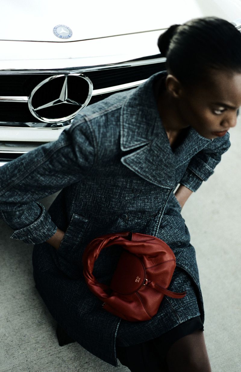 A woman in a gray trench coat stands near a white Mercedes-Benz car, accessorizing with a red leather purse in an indoor environment.