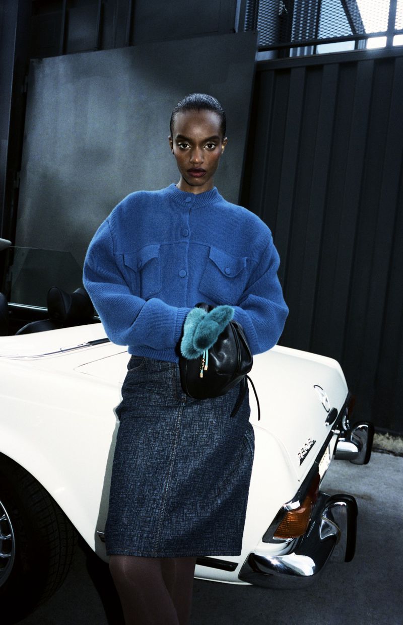 Fashion portrait of a woman in blue jacket and denim skirt posing next to a white car in a studio setting with dark backdrop.